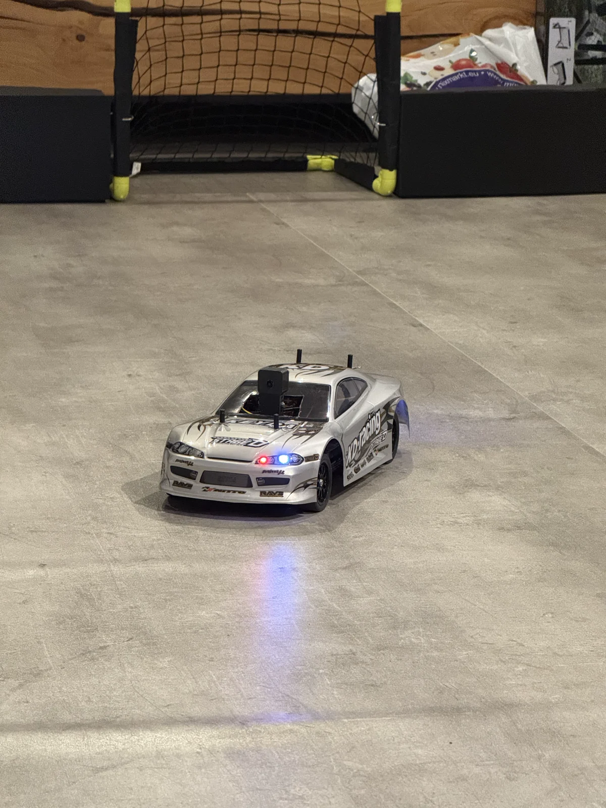 A white RC car with its lights on in front of a small goal